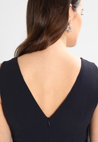 Ärmlös klänning med djup V-urtavla, tillverkad av mörkt tyg, med en dold dragkedja på baksidan och utan synliga mönster.