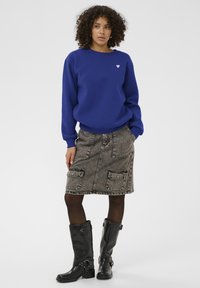 Modrá mikina s malým bílým srdíčkovým logem, doplněná o šedou džínsovou sukni s kapsami a černé vysoké boty s kovovými detaily.