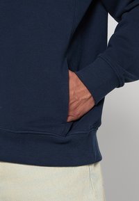 Sudadera azul marino con puños acanalados, bolsillo tipo canguro y suave tejido de mezcla de algodón. Primer plano de un bolsillo lateral con una mano insertada.