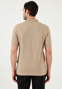 Béžové polo tričko s krátkým rukávem, s klasickým límcem, hladkou texturou a minimalistickým designem. Bez vzorů nebo viditelných log.