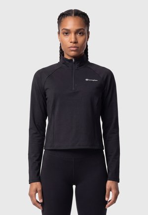 Frau mit geflochtenem Haar trägt einen schwarzen Pullover mit langen Ärmeln, einem Front-Reißverschluss und Champion-Logo auf der Brust vor einem einfarbigen Hintergrund.