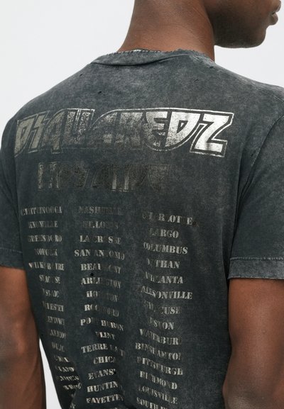 Camiseta negra descolorida con textura desgastada, que presenta un gran texto blanco y nombres de ciudades impresos en la parte posterior en varios tamaños.