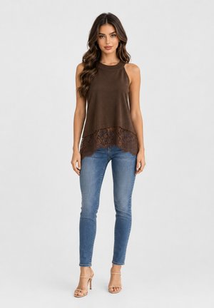 Vrouw met lang golvend haar, gekleed in een mouwloze bruine top met kanten zoom, blauwe skinny jeans en beige sandalen met hoge hakken, staand tegen een effen achtergrond.