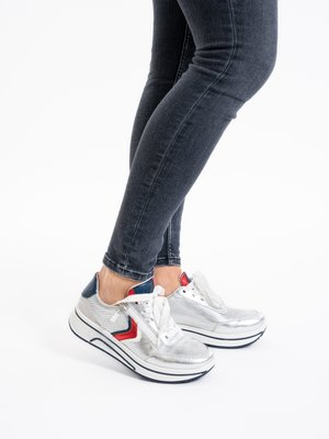 Beine in dunklen Jeans tragen silberne Plateau-Sneaker mit roten und marineblauen Akzenten, stehend vor einem weißen Hintergrund.