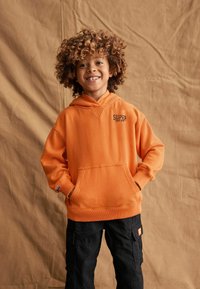Felpa arancione in cotone con tasca frontale, polsini a costine e logo sul petto. Abbinata a pantaloni cargo neri, su uno sfondo beige.
