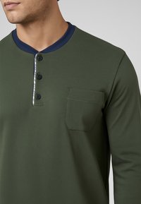 Maglietta verde oliva a maniche lunghe con colletto blu navy, tre bottoni neri e una tasca sul petto; texture liscia e design casual.