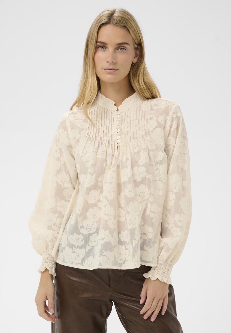 Femme portant un chemisier floral crème transparent avec des détails de plis, des boutons en perle, des poignets smockés, associé à un pantalon en cuir marron, debout devant un fond blanc.