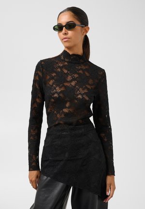 Femme portant un haut en dentelle noire à manches longues avec col montant, une jupe en dentelle asymétrique assortie, un pantalon en cuir noir et des lunettes de soleil foncées.