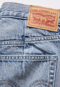 Ljusblå denimjeans med en brun läderetikett med präglade grafiska element och text som anger märke och kvalitet. Sliten textur, synlig sömnad.