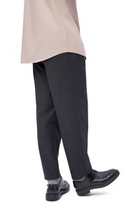 Personne portant un pantalon gris foncé, des chaussettes gris clair, des chaussures habillées noires et une chemise beige, debout sur fond blanc.