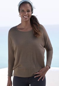 Braunes Langarmoberteil aus einem weichen, strukturierten Stoff mit rundem Ausschnitt, kombiniert mit dunkelblauen Jeans. Heller Hintergrund.