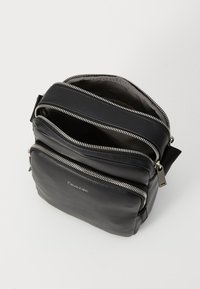 Borsa a tracolla in pelle nera con molteplici scomparti con zip e il logo Calvin Klein sulla parte anteriore, su uno sfondo neutro.