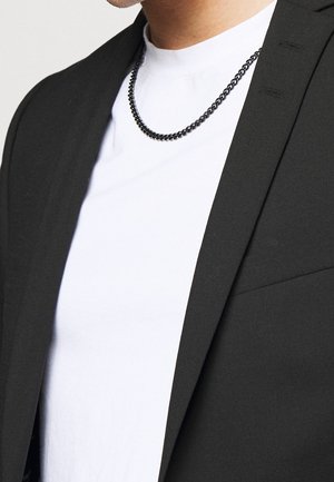 Homme portant un collier chaîne noir sur un t-shirt blanc avec un blazer noir, cadré du cou à la mi-torse.