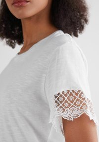 Witte, korte mouwen t-shirt met delicate kantafwerking op de mouwen, versierd met bloemenpatronen en een licht gestructureerde stof.
