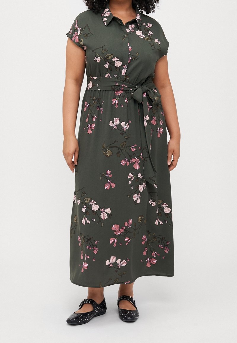 Donna che indossa un vestito maxi floreale verde scuro a maniche corte, con un nastro in vita e scarpe Mary Jane nere con borchie, su uno sfondo semplice.