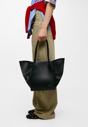 Persona che indossa pantaloni beige, camicia a righe blu, maglione rosso legato in vita, tiene una grande borsa a mano nera traforata con il logo TOUS.