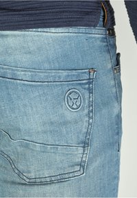 Light blue denim jeans featuring a back pocket with stitched accents and a circular logo. The fabric has a slightly worn texture.