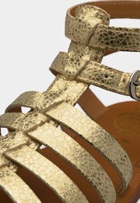 Χρυσό μεταλλικό δερμάτινο σανδάλι με πολλαπλούς ιμάντες και ασημένια πόρπη σε καφέ σόλα.