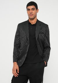 Mees, kes kannab mustrilist musta bleiserit musta polo särgi ja mustade pükstega, seisab ühe käega taskus valge tausta ees.