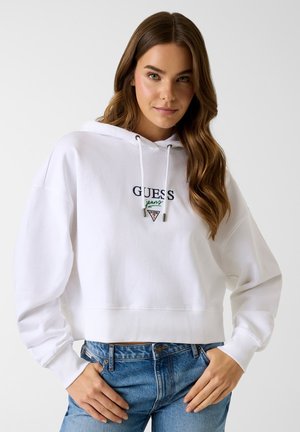 Jeune femme aux longs cheveux bruns portant un sweat à capuche blanc Guess et un jean en denim bleu, debout les mains sur les hanches devant un fond uni.