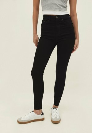 High-rise black skinny jeans with a raw hem, made from stretchy fabric, paired with white sneakers featuring grey and black accents.