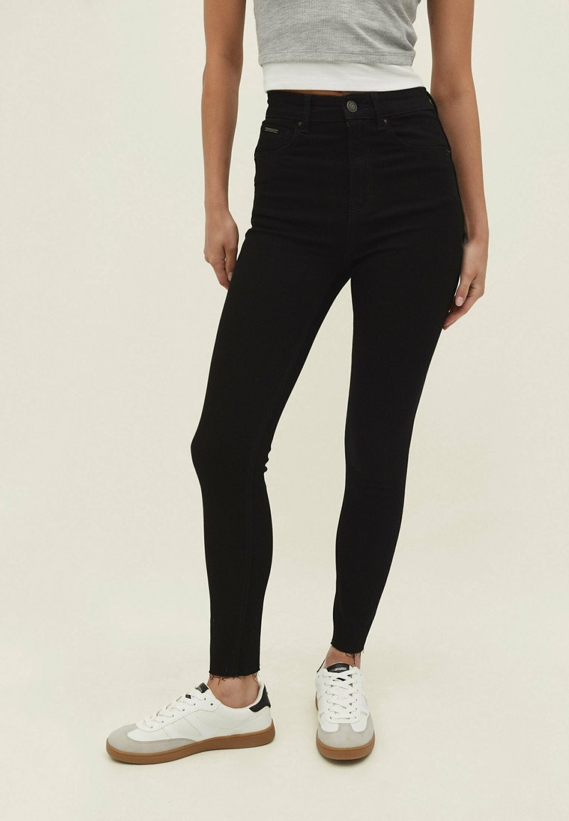 Jeans ajustados negros de cintura alta con un dobladillo sin rematar, hechos de un tejido elástico, combinados con zapatillas blancas con acentos en gris y negro.