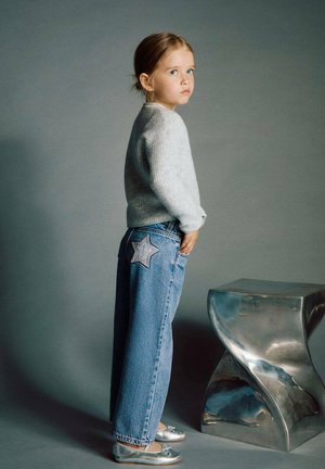 Young girl stands sideways wearing a gray sweater, blue jeans with a star patch, silver shoes, next to a reflective curved metal stool.