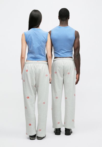 Divas personas stāv mugurām pret kameru, valkājot saskaņotas gaiši zilas piedurkņu krekliņus un pelēkas sporta bikses ar sarkanu lūpu druku rakstiem.