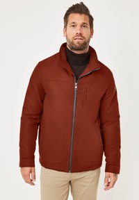 Braune gesteppte Jacke mit hohem Kragen, Frontreißverschluss und zwei seitlichen Taschen. Über einem schwarzen Rollkragenpullover getragen. Glatte Textur.