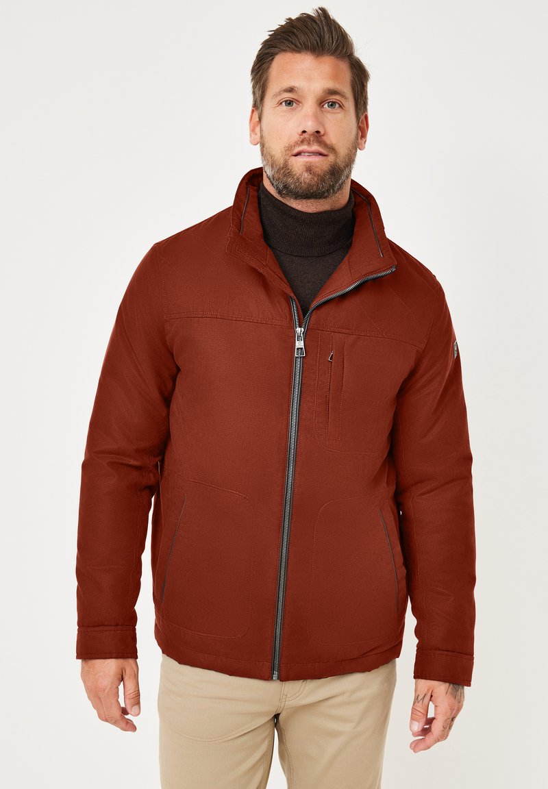 Braune gesteppte Jacke mit hohem Kragen, Frontreißverschluss und zwei seitlichen Taschen. Über einem schwarzen Rollkragenpullover getragen. Glatte Textur.