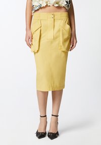 Žuta olovka suknja ima prilagođeni dizajn s bočnim džepovima i gumiranim pojasom. Tkanina je glatka, a suknja je do koljena.