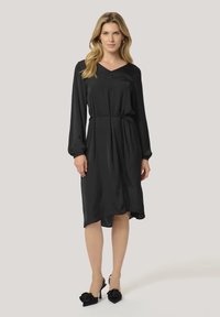 Robe longue noire à manches longues avec un décolleté en V, taille cintrée et silhouette fluide. Associée à des chaussures à talons hauts noires ornées d'accents floraux.