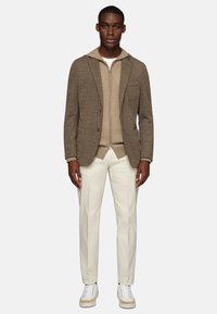 Brown textured blazer over a beige zip-up hoodie, paired with off-white trousers and white sneakers with beige accents.