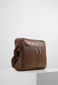 Brązowa skórzana torba na ramię z dwoma przednimi zamykanymi na zamek kieszeniami i regulowanym materiałowym paskiem na ramię, umieszczona na białym piedestale na jednolitym tle.