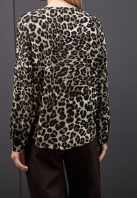 Femme portant un haut à manches longues imprimé léopard beige et noir associé à un pantalon noir, debout devant un fond gris foncé.