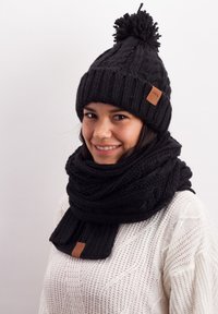Black knitted beanie with a pom-pom and a leather label, paired with a thick knitted black scarf. Both items feature textured patterns.