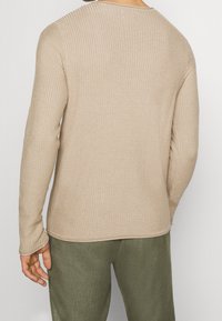 Pull beige texturé avec un col rond et des manches longues, présentant un motif côtelé et des bords finis. Associé à un pantalon vert olive.