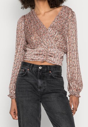 Mujer con blusa floral cruzada de manga larga con escote en V y jeans negros de cintura alta, de pie con una mano relajada.
