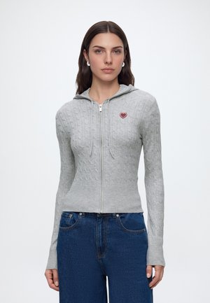 Mujer que lleva una sudadera con capucha de punto trenzado en gris claro con cremallera y un pequeño parche de corazón rojo, combinada con vaqueros azules de tiro alto.