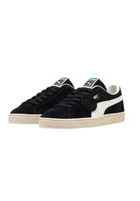 Sneaker in suede nera con una striscia bianca laterale, suola in gomma beige, chiusura con lacci, etichetta logo verde e un accento circolare sul lato.