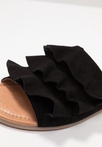 Svart mocka slide sandal med volangdetaljer, en mjuk brun fotbädd och en platt sula med en slät, matt finish.