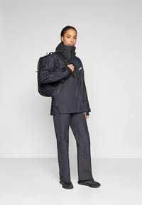 Γυναίκα φορεμένη με μαύρη εξωτερική ζακέτα και παντελόνι, κρατώντας ένα σακίδιο, που στέκεται μπροστά σε απλό φόντο.