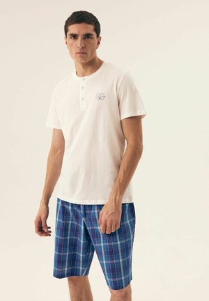 Weißes Henley-Shirt mit kurzen Ärmeln und drei Knöpfen, kombiniert mit blauen Karotten-Shorts mit grünen und lila Akzenten. Glatte Textur, lässige Passform.