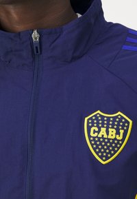 Námořnická bunda na zip s vysokým límcem, zdobená žlutým štítovým logem s modrými hvězdami a nápisem "CABJ." Texturovaná látka s modrými pruhy na ramenou.