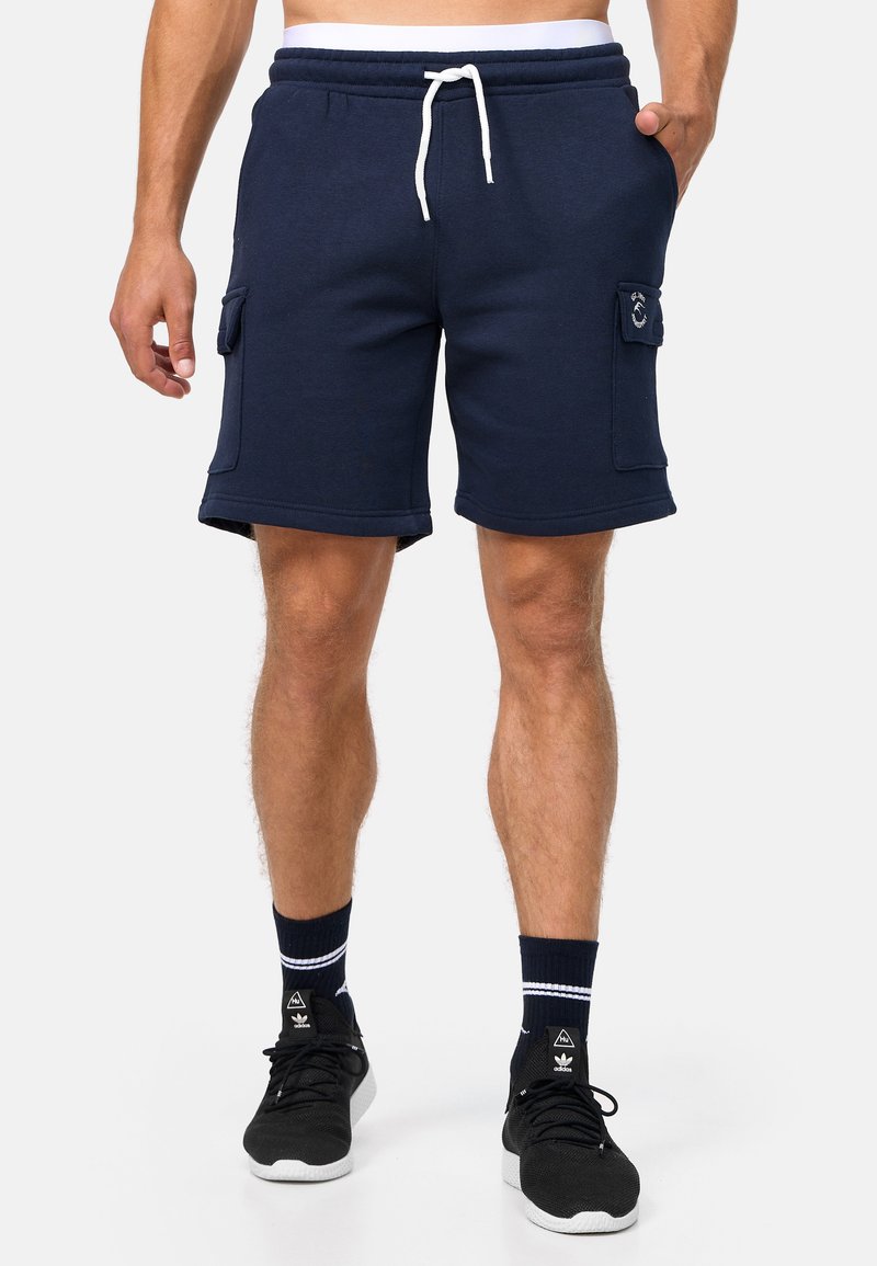 Shorts en mélange de coton bleu marine avec ceinture élastique, cordon blanc, poches latérales et coutures plates. Portés avec des baskets noires et des chaussettes navy.