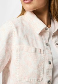 Giacca in denim maculato rosa chiaro con chiusura a bottoni, colletto a punta e tasca a toppa. Presenta bottoni metallici e una finitura strutturata.