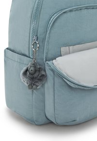 Mochila de tela azul clara con un cierre de cremallera prominente, un llavero de pelaje gris suave con un figurita y un bolsillo frontal.