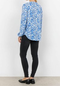 Niebieska bluzka w kwiatowy wzór z długimi rękawami, luźny krój i zaokrąglony dół, zestawiona z czarnymi dopasowanymi spodniami i płaskimi czarnymi butami.