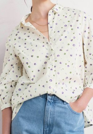 Kobieta w kremowej bluzce w zielone i fioletowe groszki, włożonej w jasnoniebieskie dżinsy, z ręką w kieszeni, nosząca warstwowe złote naszyjniki.