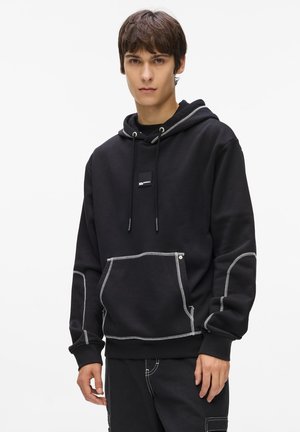 Hoodie noir en tissu mélangé coton, avec une poche devant, des coutures blanches contrastantes et une capuche avec cordon de serrage.